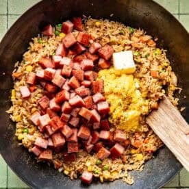 A wok containing fried rice, diced spam, scrambled eggs, vegetables, and a pat of butter, with a wooden spatula resting on the side.