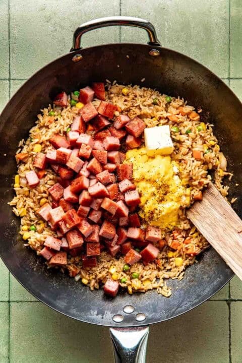 A wok containing fried rice, diced spam, scrambled eggs, vegetables, and a pat of butter, with a wooden spatula resting on the side.