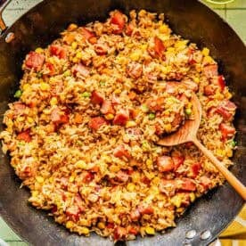 A skillet filled with fried rice mixed with diced ham, corn, peas, and carrots, with a wooden spoon resting inside.