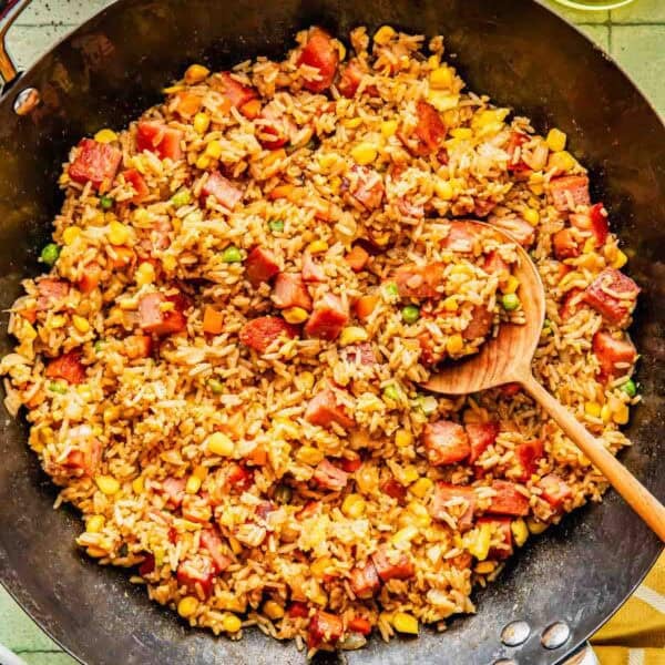 A skillet filled with fried rice mixed with diced ham, corn, peas, and carrots, with a wooden spoon resting inside.