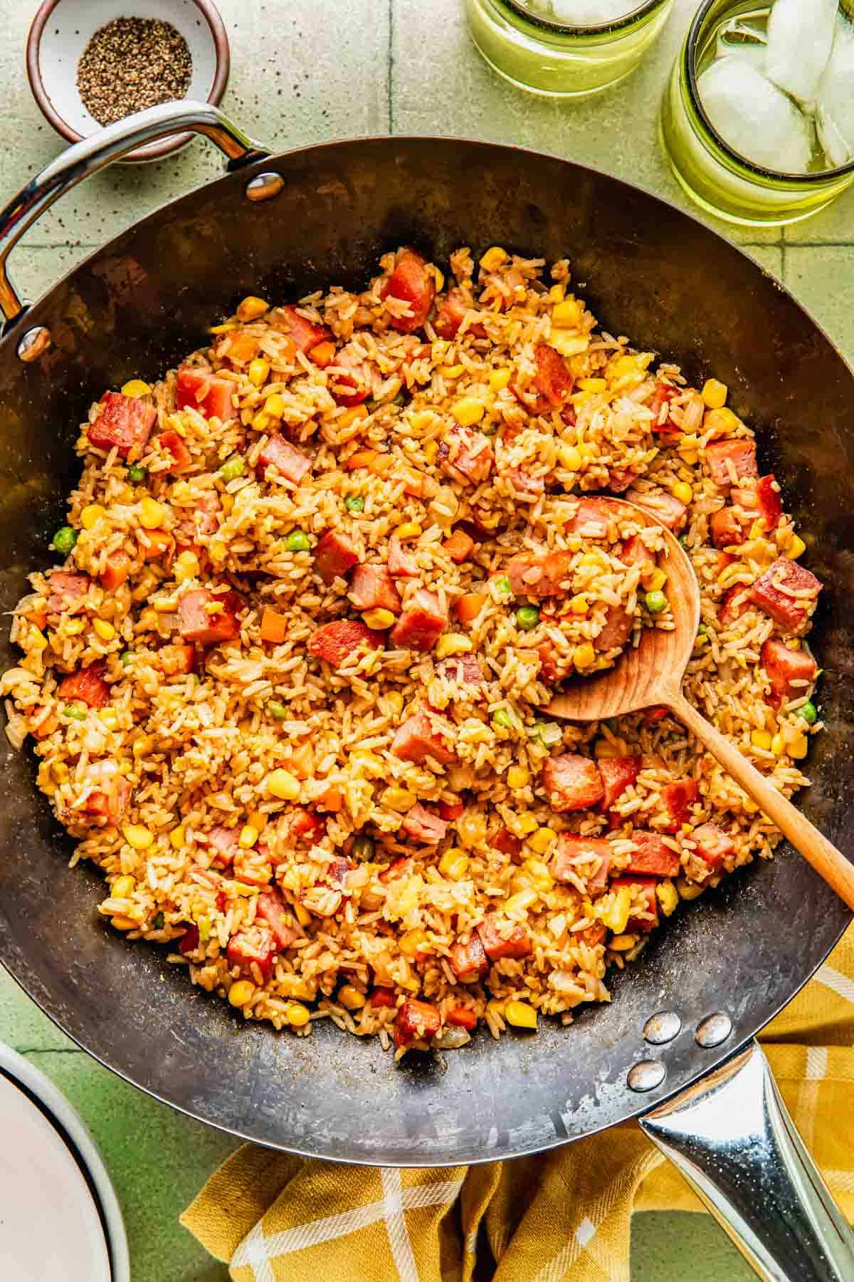 A skillet filled with fried rice mixed with diced ham, corn, peas, and carrots, with a wooden spoon resting inside.
