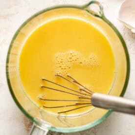 Clear glass bowl with yellow egg mixture being whisked, with eggshells visible in the background on a light surface.