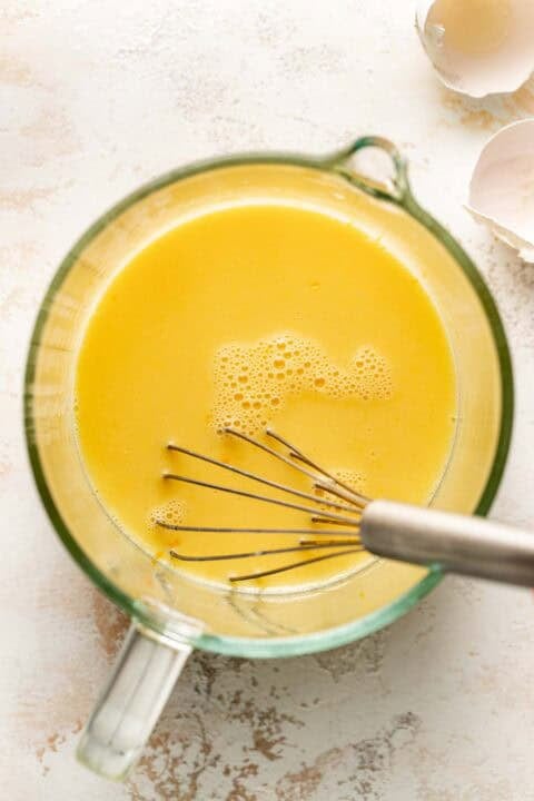 Clear glass bowl with yellow egg mixture being whisked, with eggshells visible in the background on a light surface.