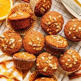 Twelve oat-topped muffins are arranged on a cooling rack, with a few unwrapped; nearby are orange slices, oats, cinnamon sticks, butter, and a yellow-striped napkin.