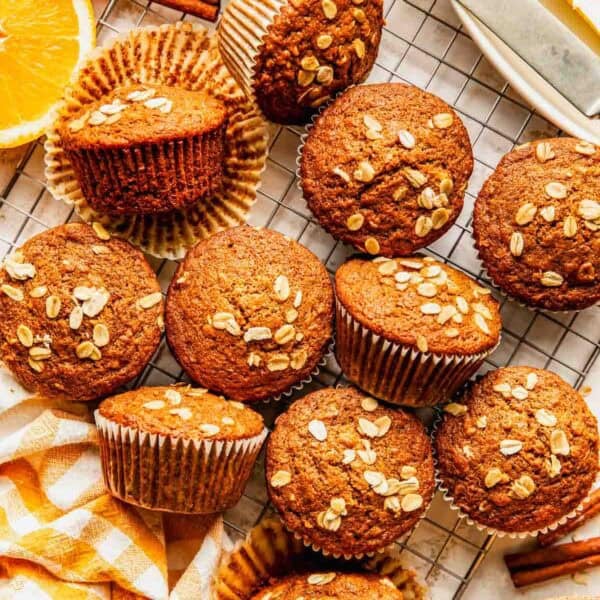 Twelve oat-topped muffins are arranged on a cooling rack, with a few unwrapped; nearby are orange slices, oats, cinnamon sticks, butter, and a yellow-striped napkin.