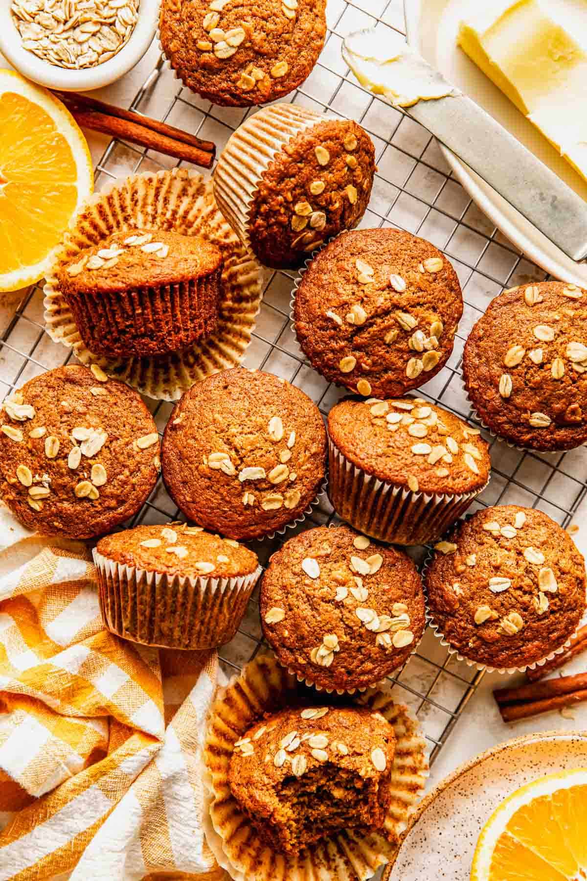 Twelve oat-topped muffins are arranged on a cooling rack, with a few unwrapped; nearby are orange slices, oats, cinnamon sticks, butter, and a yellow-striped napkin.