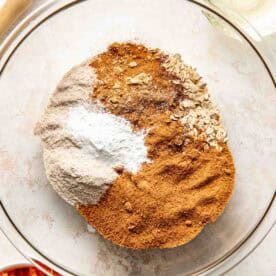 A glass bowl with flour, oats, brown sugar, baking powder, cinnamon, and other dry ingredients; nearby are shredded carrots, a whisk, and a small bowl of oil.