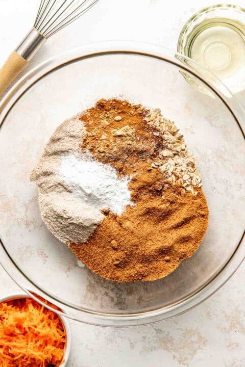A glass bowl with flour, oats, brown sugar, baking powder, cinnamon, and other dry ingredients; nearby are shredded carrots, a whisk, and a small bowl of oil.