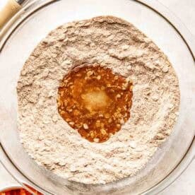 A glass mixing bowl with flour and oats, forming a well in the center filled with liquid ingredients; a whisk and grated carrots are nearby.