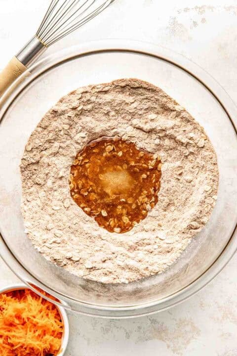 A glass mixing bowl with flour and oats, forming a well in the center filled with liquid ingredients; a whisk and grated carrots are nearby.