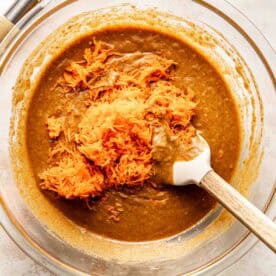 A glass bowl of brown batter with shredded carrots being mixed in with a spatula; a whisk is nearby on the counter.