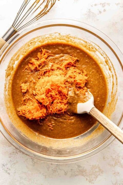 A glass bowl of brown batter with shredded carrots being mixed in with a spatula; a whisk is nearby on the counter.