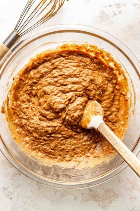 A glass bowl filled with brown muffin batter being mixed with a rubber spatula, with a whisk lying nearby on a white surface.