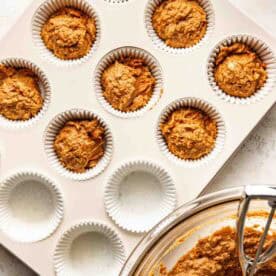 Muffin tin with paper liners, some filled with unbaked muffin batter. A glass bowl with more batter and a scoop are next to the tin.