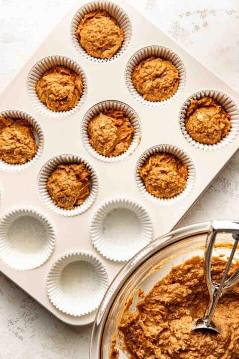 Muffin tin with paper liners, some filled with unbaked muffin batter. A glass bowl with more batter and a scoop are next to the tin.