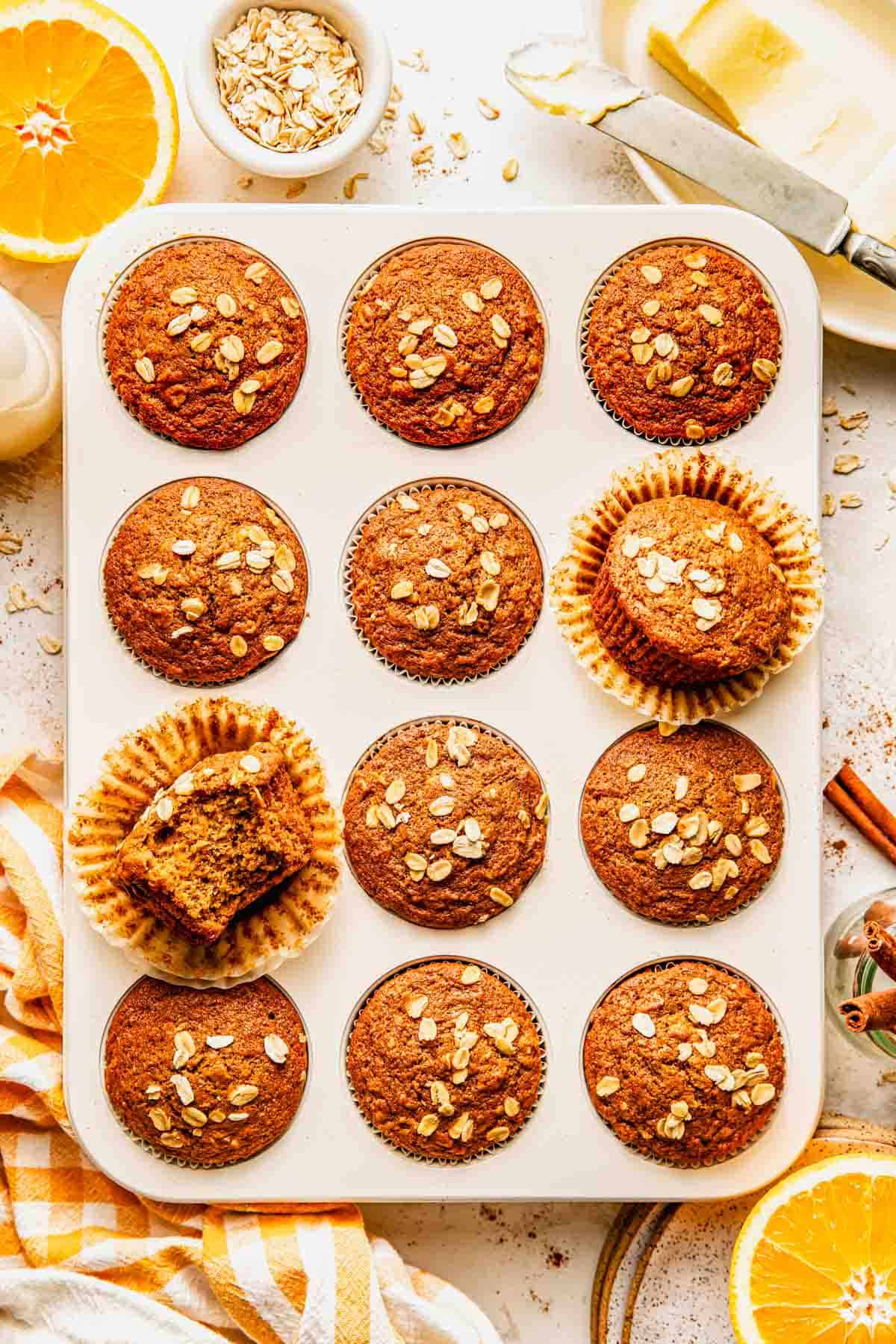 A muffin tin filled with twelve baked oat muffins, some with paper liners, one partially unwrapped and one with a bite taken out. Sliced oranges, oats, butter, and cinnamon are nearby.