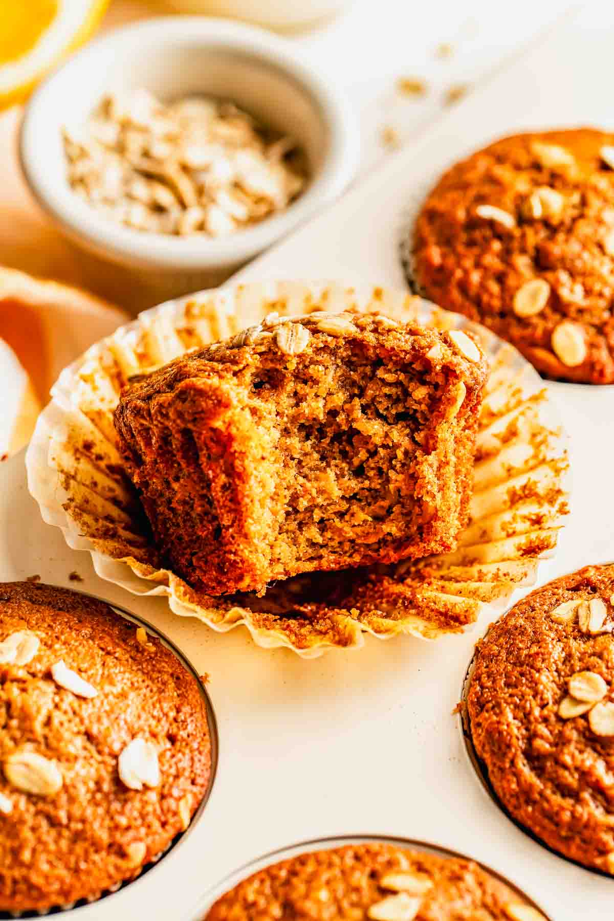 A close-up of an oat muffin with a bite taken out, sitting in a paper liner among other whole muffins, with oats sprinkled on top.