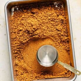 A metal baking pan with crushed graham crackers and a 1-cup metal measuring cup resting on top of the crumbs.