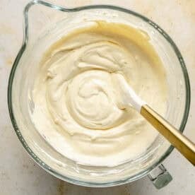 A glass mixing bowl filled with creamy batter being stirred with a white and gold spatula on a light countertop.