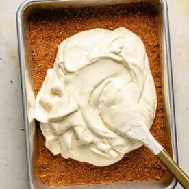 A spatula spreads cheesecake filling over a graham cracker crust in a rectangular metal baking pan.