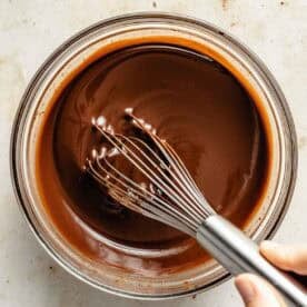 A hand holds a metal whisk stirring smooth melted chocolate in a clear glass bowl on a light-colored surface.