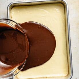 A glass bowl pours melted chocolate onto a rectangular pan of baked cheesecake, creating a chocolate layer on top.