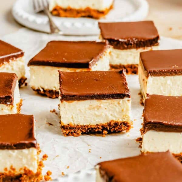 Nine cheesecake bars with a graham cracker crust and chocolate topping are arranged on parchment paper and a white plate.