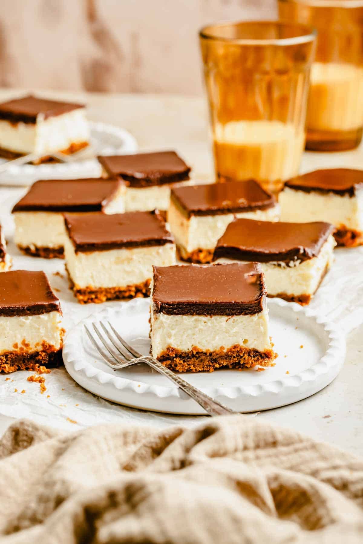 Squares of cheesecake with a chocolate topping and graham cracker crust are arranged on white plates, accompanied by a fork and glasses of amber-colored liquid in the background.