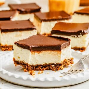 A square cheesecake bar with a chocolate topping and crumbly crust sits on a plate with a fork, surrounded by similar bars in the background.