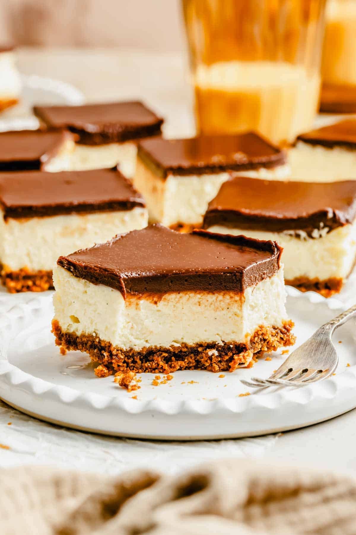 A square cheesecake bar with a chocolate topping and crumbly crust sits on a plate with a fork, surrounded by similar bars in the background.