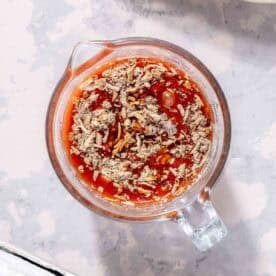 A glass measuring cup filled with a mixture of tomato sauce and dry seasoning on a light countertop.