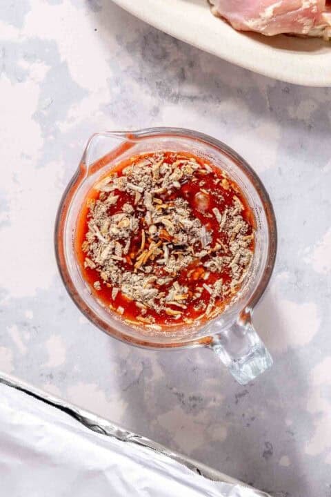 A glass measuring cup filled with a mixture of tomato sauce and dry seasoning on a light countertop.