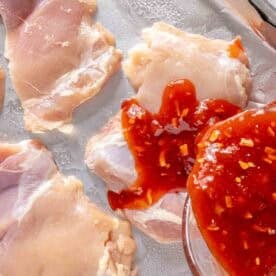 Raw chicken thighs on a baking sheet are being topped with red sauce from a glass bowl.