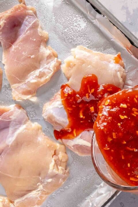 Raw chicken thighs on a baking sheet are being topped with red sauce from a glass bowl.