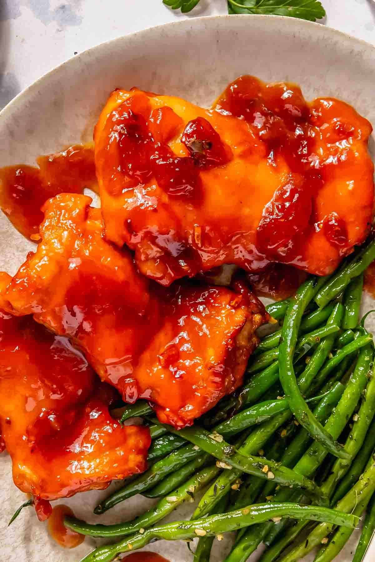 Plate with three pieces of glazed chicken topped with red sauce, served alongside sautéed green beans on a white dish.