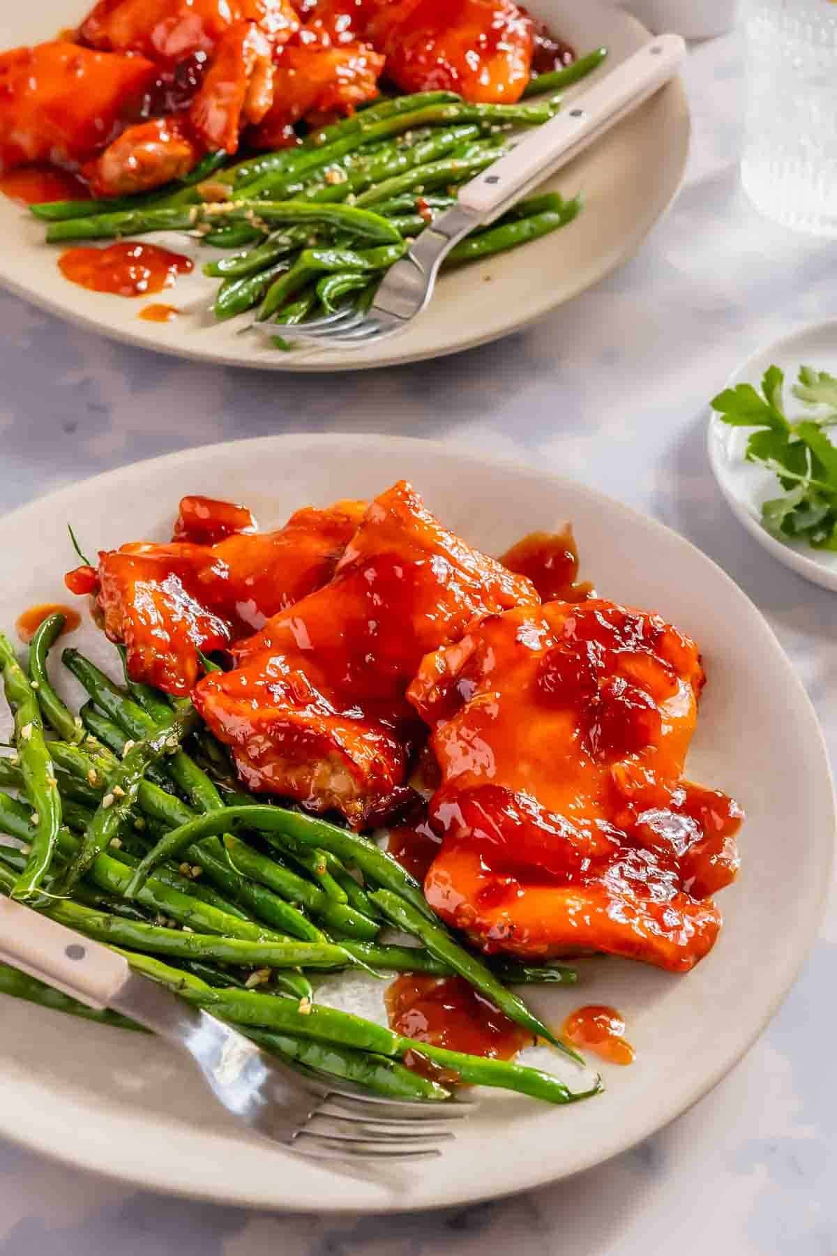 Plate of cooked green beans and glazed chicken with red sauce, served with a fork on a beige plate. Another similar plate and a small dish of herbs are in the background.