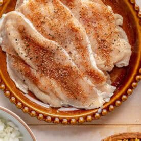 Seasoned raw chicken breasts on a brown plate, with bowls of chopped onions and uncooked rotini pasta nearby.