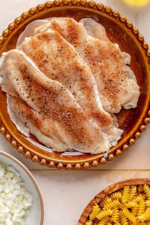 Seasoned raw chicken breasts on a brown plate, with bowls of chopped onions and uncooked rotini pasta nearby.