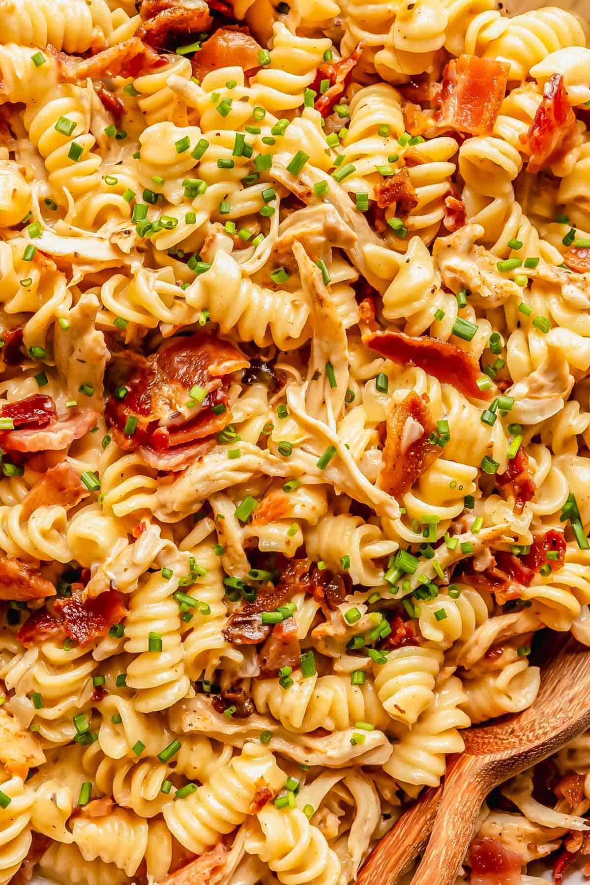 Rotini pasta with shredded chicken, crispy bacon, creamy sauce, and chopped chives, served with wooden utensils.