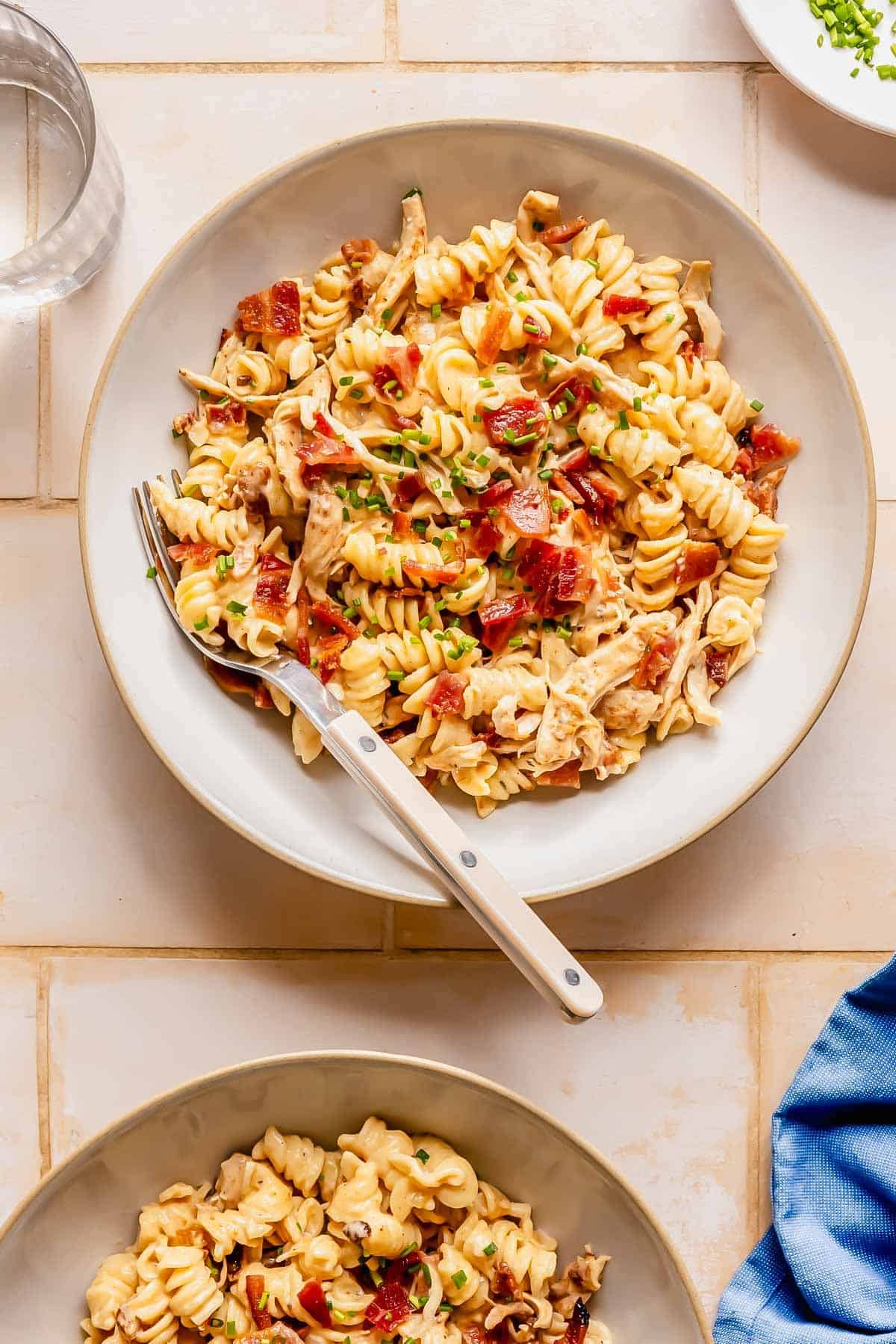 Bowl of rotini pasta mixed with shredded chicken, bacon pieces, and chopped herbs, served with a fork on a light tiled surface.