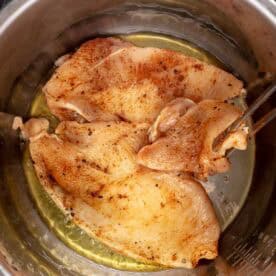 Seasoned chicken breasts cooking in oil inside a stainless steel pressure cooker, with tongs holding one piece.