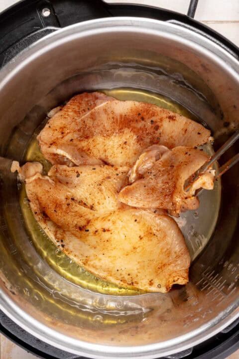 Seasoned chicken breasts cooking in oil inside a stainless steel pressure cooker, with tongs holding one piece.