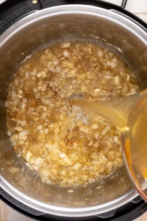 Chopped onions and garlic are sautéed in an Instant Pot, while broth is being poured in, viewed from above.