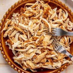 A brown plate filled with shredded, seasoned chicken sits on a tiled surface with two forks resting on top.