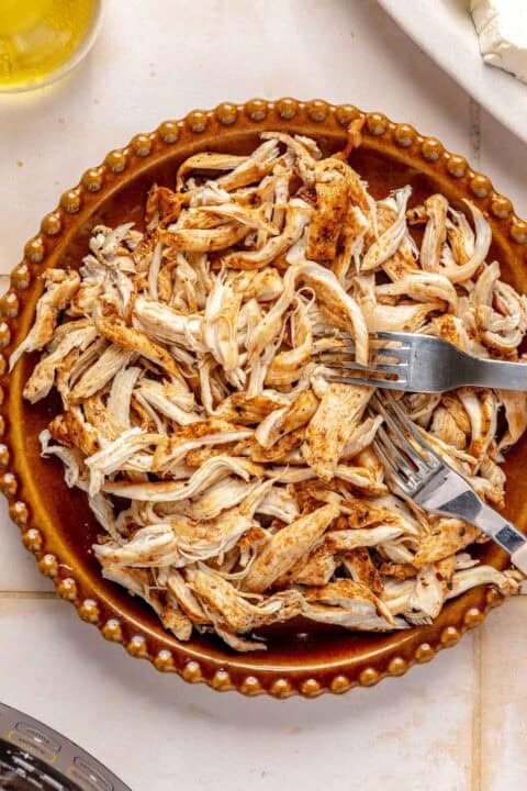 A brown plate filled with shredded, seasoned chicken sits on a tiled surface with two forks resting on top.