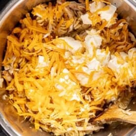 Shredded cheese and cream cheese on top of cooked pasta in an Instant Pot, with a wooden spoon partially visible.