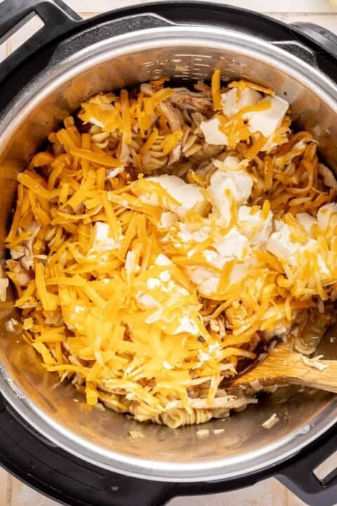 Shredded cheese and cream cheese on top of cooked pasta in an Instant Pot, with a wooden spoon partially visible.