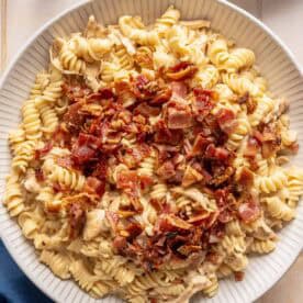 A plate of rotini pasta topped with chopped cooked bacon and pieces of chicken, set on a light-colored table next to a blue napkin, glass of water, and white plates.