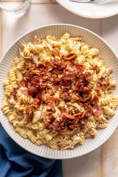 A plate of rotini pasta topped with chopped cooked bacon and pieces of chicken, set on a light-colored table next to a blue napkin, glass of water, and white plates.