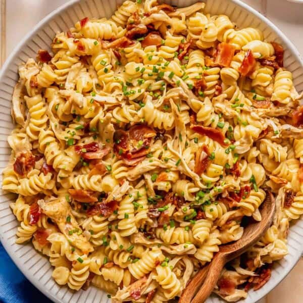 A plate of rotini pasta mixed with shredded chicken, crispy bacon, and chopped chives, with a wooden spoon resting in the dish.